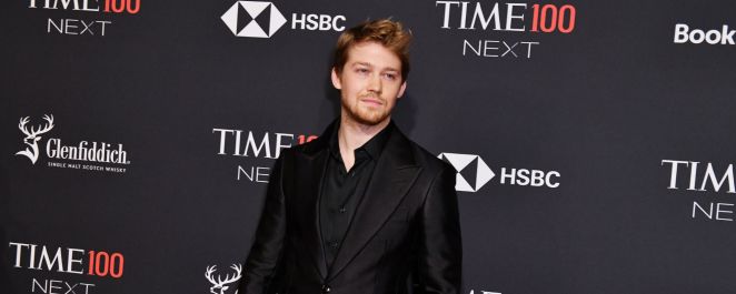 Joe Alwyn, Taylor Swift's ex-boyfriend, wears a black suit standing against a black background with white writing on it.