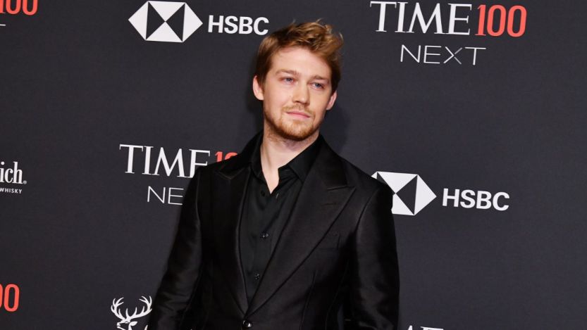 Joe Alwyn, Taylor Swift's ex-boyfriend, wears a black suit standing against a black background with white writing on it.