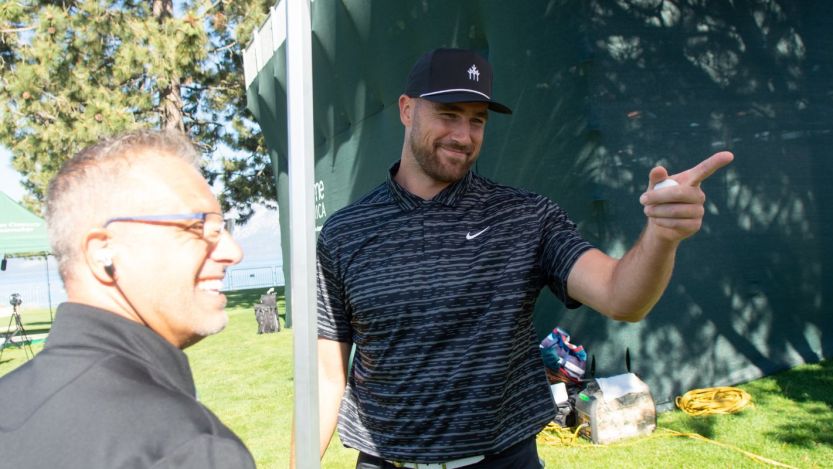 Travis Kelce wears a blue Nike shirt and hat as he points to someone off camera.
