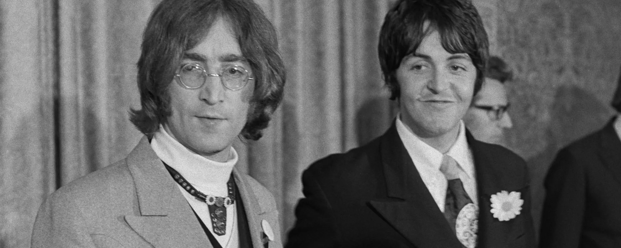 John Lennon looks off to the side, Paul McCartney stands beside him smiling