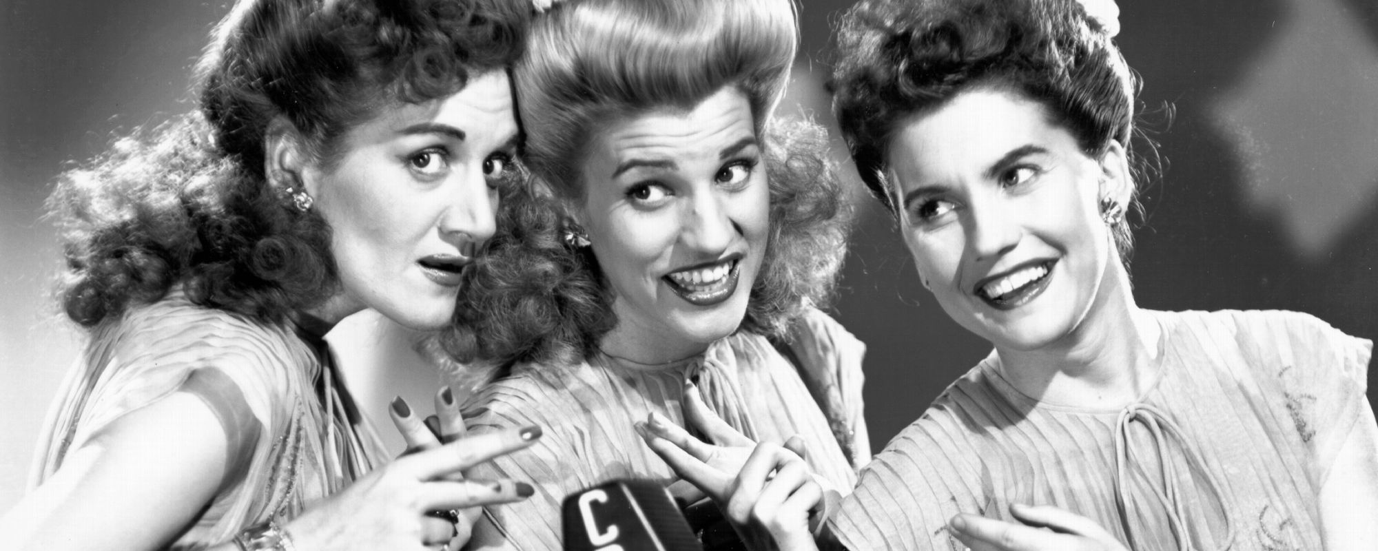 Andrews Sisters, group who sang location-specific Christmas song "Mele Kelikimaka," posing around mic