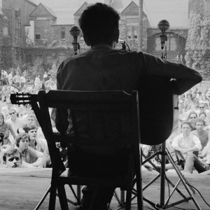 View, from behind, of American musician Bob Dylan as he plays acoustic guitar during a performance at the Newport Folk Festival, Newport, Rhode Island, July 1963. Audience members sit on the grass of the tennis court at the ivy-covered buildings of the Newport Casino (later the National, and then International, Tennis Hall of Fame). (Photo by Rowland Scherman/Getty Images)
