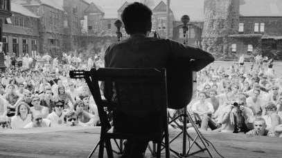 View, from behind, of American musician Bob Dylan as he plays acoustic guitar during a performance at the Newport Folk Festival, Newport, Rhode Island, July 1963. Audience members sit on the grass of the tennis court at the ivy-covered buildings of the Newport Casino (later the National, and then International, Tennis Hall of Fame). (Photo by Rowland Scherman/Getty Images)